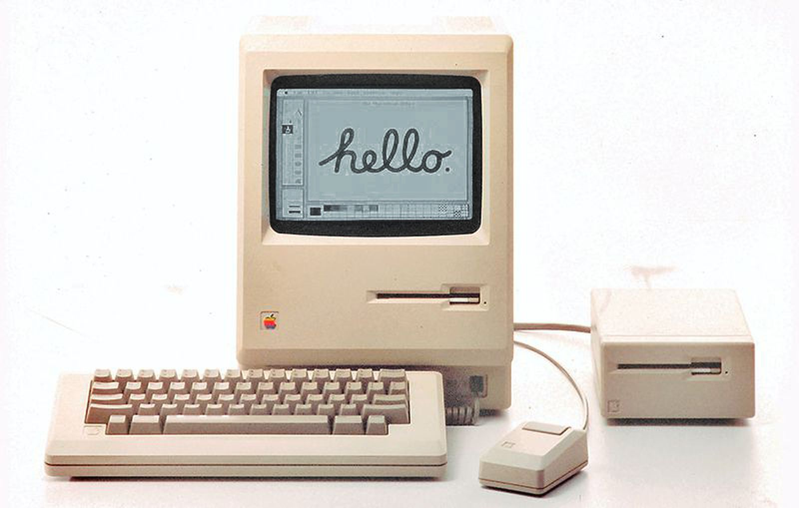 Publicity shot of Apple's original 128K Mac, an upright rectange in Apple's trademark beige with a built-in black-and-white screen. Attached, a keyboard, mouse, and external floppy drive.