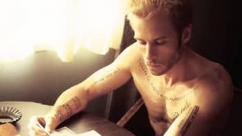 Leonard Shelby, a character in Christopher Nolan's film Memento, sits shirtless and tattooed at a hotel tabel.