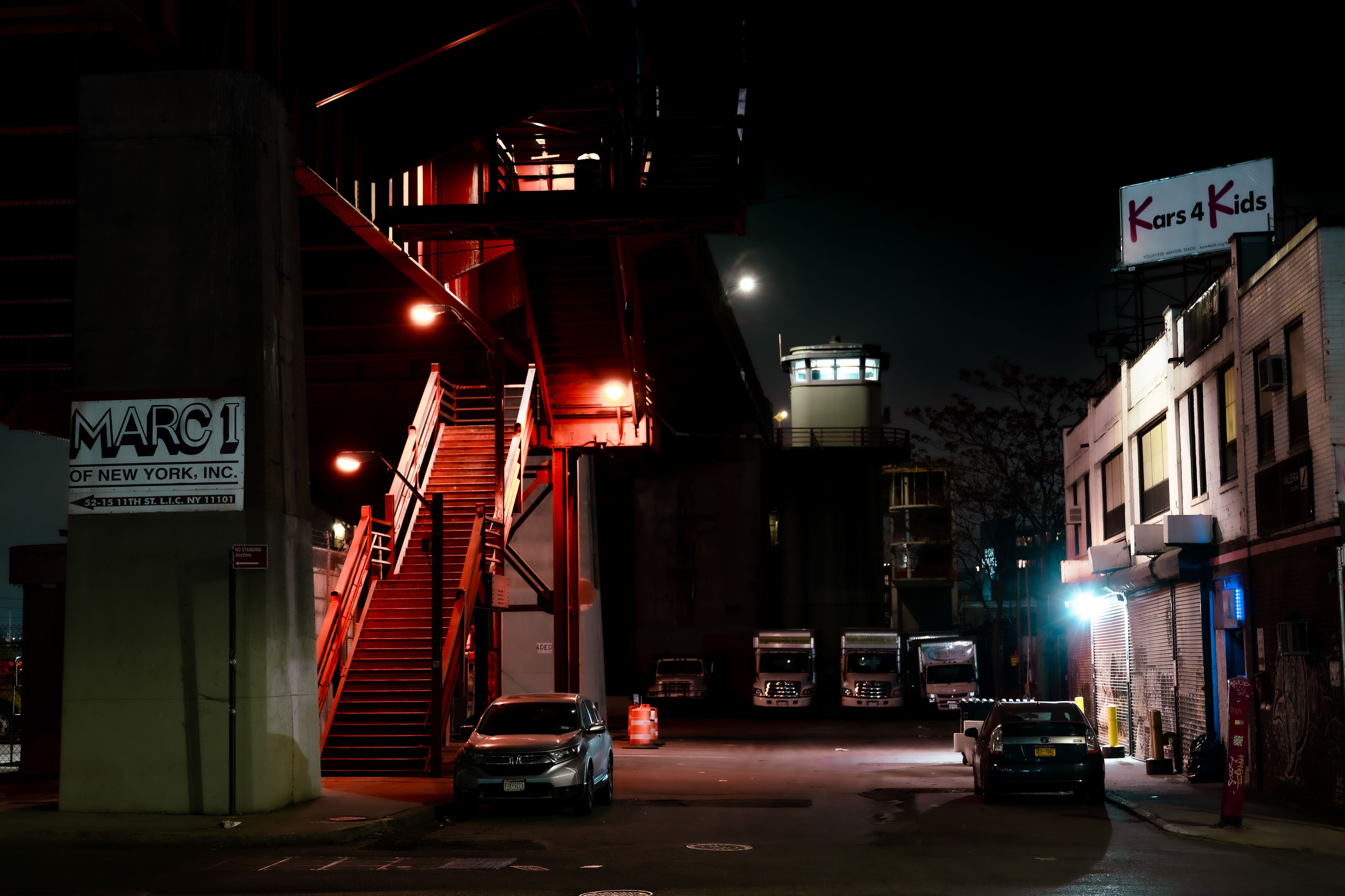 Dark streetscape of a city lot with red stairwell up to a bridge.